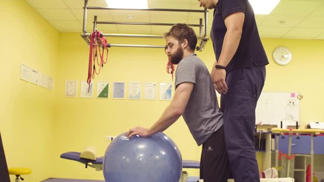 Young disabled man doing exercises at the rehabilitation center with a ball. Doctor physiotherapist helping him. Healthy gymnastics. Active people.