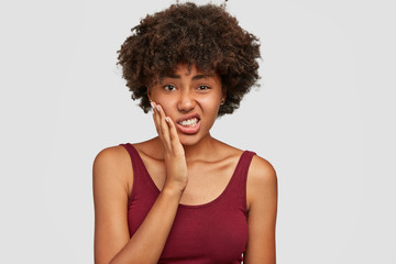 Fototapeta na wymiar Displeased beautiful African American woman keeps hand on cheeks, feels toothache, clenches teeth from pain, has dark healthy skin and crisp hair, dressed casually. Negative facial expressions