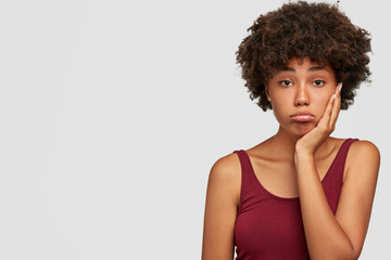 Upset beautiful African American female keeps hand on cheeks, looks displeased after parting with boyfriend, purses lower lip, dressed in casual red t shirt, isolated on white studio wall, blank space
