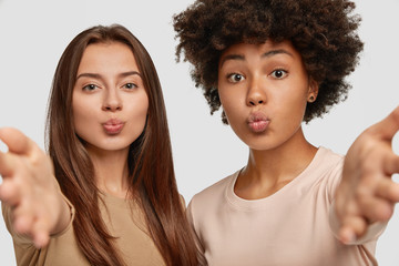 Lovely mixed race women pout lips, stretch hands at camera as going to embrace someone, express their good feeling, pose together against white background. Interracial female friends indoor.