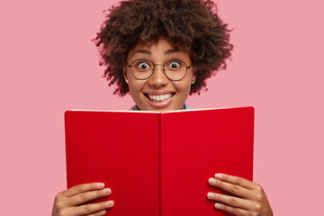 Horizontal shot of happy African American female holds red book, has positive expression, reads something interesting, isolated over pink background. Lovely dark skinned student prepares for classes