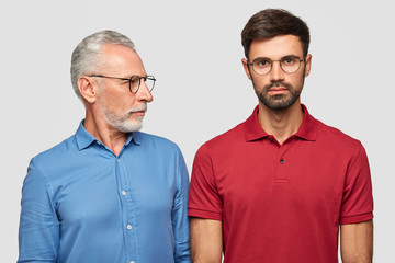 Elderly experienced male looks attentively at his adult son, gives pieces of advice, wears spectacles and formal blue shirt, have good relations. People, age, lifestyle and life experience concept