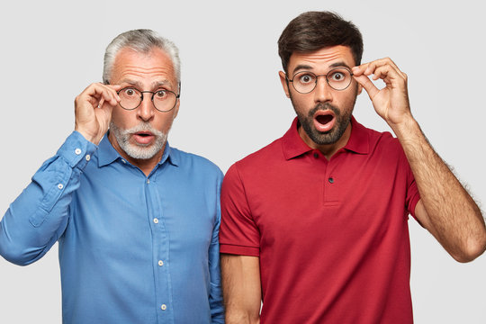 Two handsome father and sun notice something unbelievable, stare through round spectacles, dressed in bright stylish clothes, isolated over white background. Surprised bearded mature and adult men