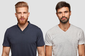 Fototapeta na wymiar Two bearded brothers in casual t shirts, have serious expressions, stand next to each other, isolated over white background. Handsome ginger hipster guy and his companion think about something
