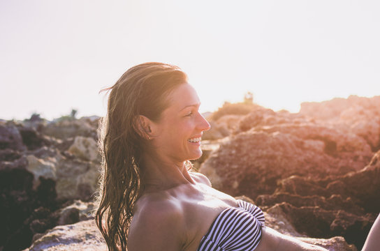 Young smiling woman with wet hair sunbathing on the rocks at sunset in bright sunshine