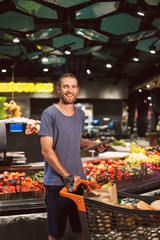 Young smiling man with trolley full of products joyfully looking in camera holding the tablet in hand in modern supermarket