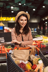 Beautiful smiling girl in hat leaning on trolley full of products joyfully looking in camera while spending time in modern supermarket