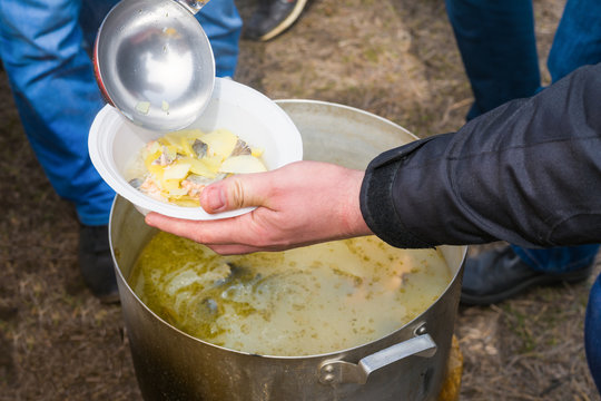 The man pours soup into a disposable plate from a large saucepan outdoor. Refugee camp, outdoor feeding, food distribution concept. 
