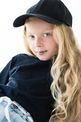 portrait of beautiful child with long curly hair wearing black t-shirt and cap, looking at camera isolated on white
