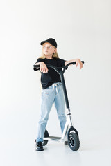 full length view of kid with long curly hair standing with scooter and looking away isolated on white