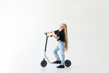 full length view of kid with long curly hair riding scooter and looking at camera isolated on white