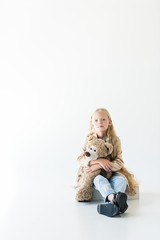 full length view of beautiful little child holding teddy bear and smiling at camera while sitting isolated on white