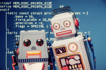 Two tin toy robots standing still like in a selfie shot. Background: computer source code (public...