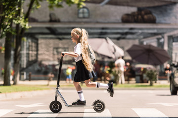 side view of cute little child with long curly hair riding scooter on street