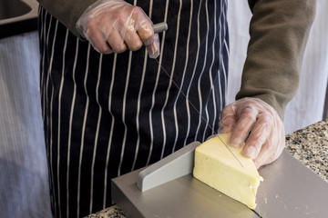 High angle close up of man wearing apron standing at counter in a delicatessen, slicing cheese with wire cutter.