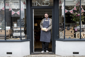 Bearded man wearing flat cap and apron standing in doorway of a delicatessen, holding cheese.