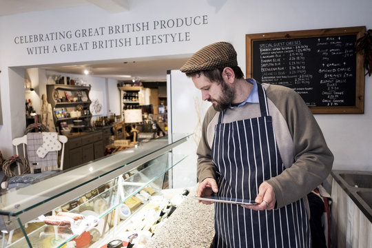 Bearded man wearing flat cap and apron standing at counter in a delicatessen, holding digital tablet. - Powered by Adobe
