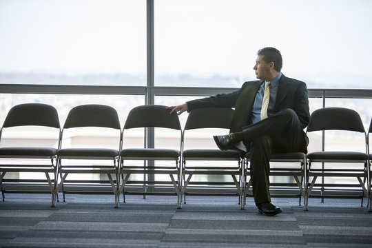 Hispanic businessman sitting in a row of chairs against a window in a conference centre lobby.