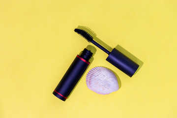 Black mascara bottle, curve brush and seashell isolated yellow background. Fashionable cosmetics.