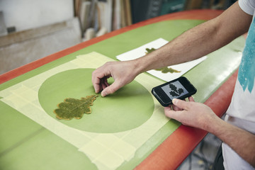Man creating a design on surfboard laying an oak leaf on the board, using mobile phone in workshop
