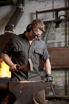 An artisan metal worker in ear protectors using a hammer to shape a red hot piece of metal on an anvil. 