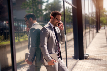 Lucky day at work.Smiling confident businessman talking with his colleague about successful ending job. Standing outside leaned on building wall.