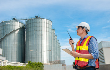 Young engineer wear hard hat using walkie talkie and tablet digital pose at set of storage tanks raw material agricultural crops feed mills. industry background