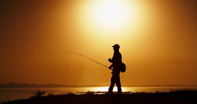 An experienced fisherman catching fish with professional spinning rod from a lake bank. lake in orange sunset 4k