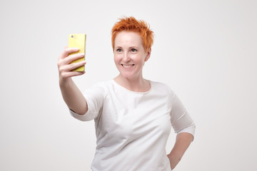 Pretty mature happy woman with red hair making selfie on smartphone