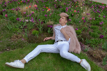 Young sexy man in a white clothes and pink coat poses in a park with flowers. Fashion concept