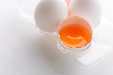 Broken egg with whole eggs in a transparent plastic container on white wooden background