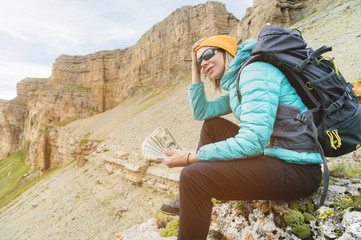 Fototapeta na wymiar A traveler girl wearing a hat and sunglasses holds a hundred dollar bills in the hands of a fan against the backdrop of cliffs on nature. Keep your head from high cost