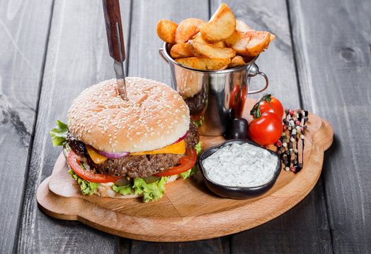 Hamburger with fresh vegetables, cheese, sauce and fries on cutting board on dark wooden background. Burger with knife