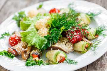 Grilled eggplant rolls and zucchini rolls with tomatoes, mushrooms, herbs, cheese balls and sesame on plate on rustic table