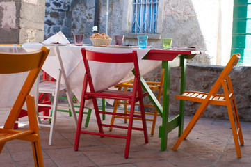 Exterior of a restaurant with a laid out table ready for lunch, with colorful chairs of red, white and natural wood color
