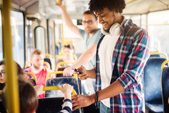 Young Afro-American With Headset Around His Neck Is Looking At His Phone While Riding In A Bus.