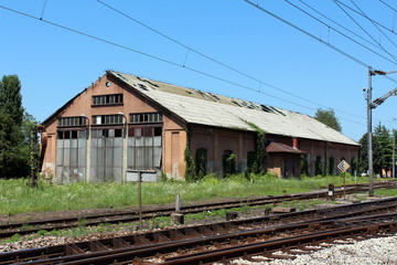Fototapeta na wymiar Dilapidated abandoned railway station building with missing roof tiles and windows surrounded with tall uncut grass and other vegetation next to rusted railway tracks with clear blue sky in background