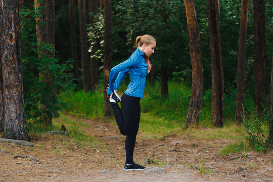 Quad stretch before run. Girl in blue sportswear doing exercises outdoors in coniferous forest. Healthy lifestyle sport concept.