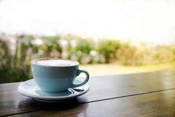 A blue cup of hot coffee on the wooden table with garden background with the sunlight from the sky with copy space. Start the good day with good coffee, hope; new day, new life concept.