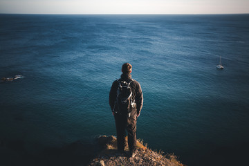 Fototapeta na wymiar A rear view of a male backpacker or hiker standing on a high clifftop and overlooking a vast ocean at Kynance Cove in Cornwall, UK