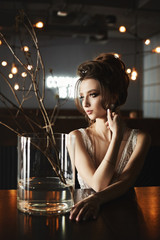 Naklejka na ściany i meble Sexy, busty and fashionable brunette model girl with bright makeup, in stylish dress sits at the table and posing at interior