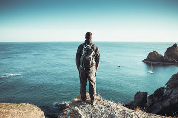 A rear view of a male backpacker or hiker standing on a high clifftop and overlooking a vast ocean...