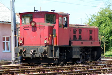 Fototapeta na wymiar Old used dark red electric locomotive parked on railway tracks waiting for departure from local railway station with trees and blue sky in background