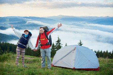 Fototapeta premium Happy couple with backpacks near the tent holding hands against the backdrop of the beautiful scenery of the mighty mountains of the Carpathians on which lies a haze and the sky is beautiful clouds.