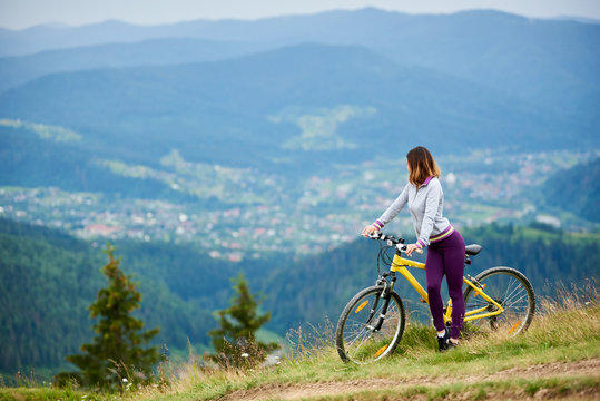 Athlete young woman cyclist with her yellow bicycle on a rural trail enjoying evening view of mountains, forests and small city on the blurred background. Outdoor sport activity. Copy space