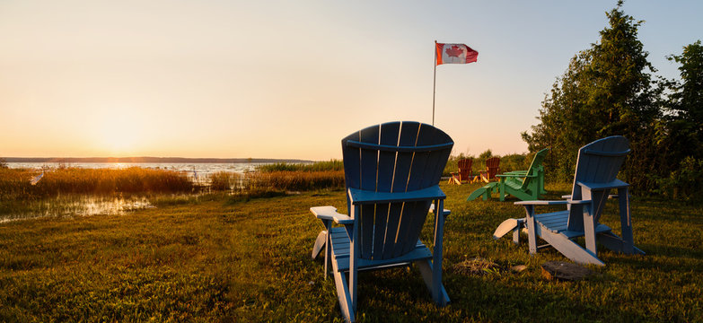 Muskoka chairs on a lawn beside a lake with a Canadian flag in the background at sunset.