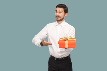 Funny happy bearded man in white shirt standing and holding red gift box, showing and looking at...