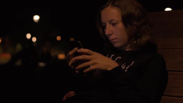 Young attractive lady sitting in the night park and wathing social media feed on her brand new mobile phone.