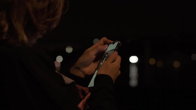 Young attractive lady sitting in the night park and wathing social media feed on her brand new mobile phone.
