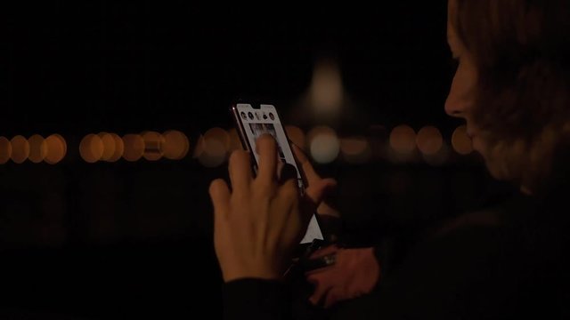 Young attractive lady sitting in the night park and wathing social media feed on her brand new mobile phone.